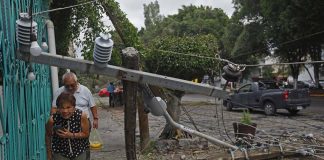 A man and woman in single file walking through debris and ducking under a downed power line on a sidewalk next to a residence on a city street. The man at the back is carrying a plastic shopping bag of bananas.