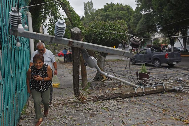 A man and woman in single file walking through debris and ducking under a downed power line on a sidewalk next to a residence on a city street. The man at the back is carrying a plastic shopping bag of bananas.