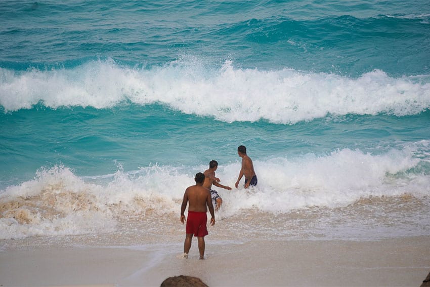 Three young men standing in the water on the shore of a Cancun beach as the waves crash on them.