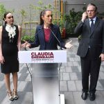 Claudia Sheinbaum standing at a podium with her name on it with her hands out before her, palms up, as she addresses reporters outside in a courtyard. To either side of her are Altagracia Gomez Sierra and Marcelo Ebrard, looking on.