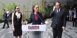 Claudia Sheinbaum standing at a podium with her name on it with her hands out before her, palms up, as she addresses reporters outside in a courtyard. To either side of her are Altagracia Gomez Sierra and Marcelo Ebrard, looking on.