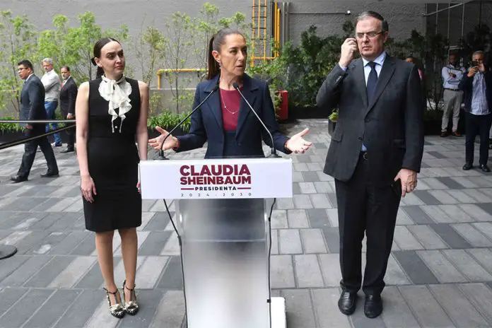 Claudia Sheinbaum standing at a podium with her name on it with her hands out before her, palms up, as she addresses reporters outside in a courtyard. To either side of her are Altagracia Gomez Sierra and Marcelo Ebrard, looking on.