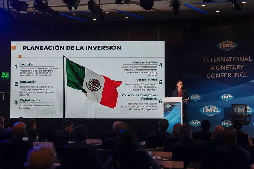 Claudia Sheinbaum at the Internationnal Monetary Conference at a podium in front of a screen projecting a six-step investment plan for her incoming government.