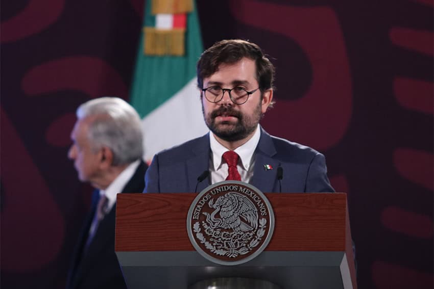 Mexico's Cofepris head Alejandro Svarch Perez standing at a podium in foreground, with Mexico's President Lopez Obrador's profile in the blurred background.