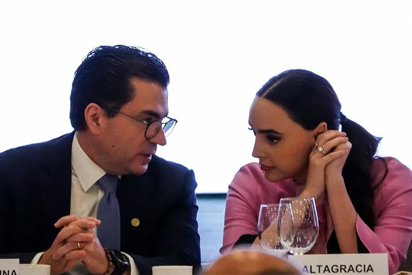 A man and a woman sitting side by side at a conference banquet table bent toward each other in conversation.