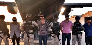 Men in handcuffs in a horizontal line in front of a military passenger jet standing among Mexican soldiers