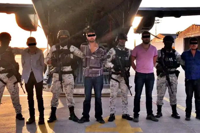 Men in handcuffs in a horizontal line in front of a military passenger jet standing among Mexican soldiers