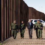 U.S. President Joe Biden walking with US Border Patrol officers along the U.S. border wall with a border patrol SUV parked in the background.