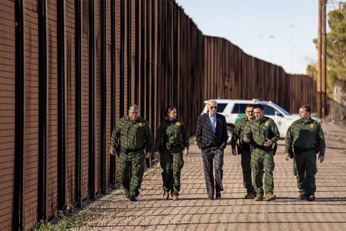 U.S. President Joe Biden walking with US Border Patrol officers along the U.S. border wall with a border patrol SUV parked in the background.