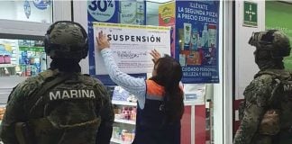 A woman putting up a sign on a pharmacy's glass door while being watched over by the Mexican navy. The sign says "Suspension"