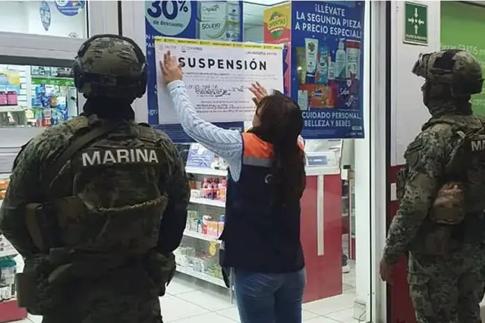 A woman putting up a sign on a pharmacy's glass door while being watched over by the Mexican navy. The sign says 