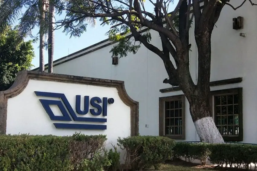 Cartel de USI en la planta de la empresa en Guadalajara, junto a un edificio de adobe y un gran árbol.