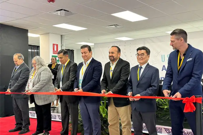 USI officials and officials from Jalisco, Mexico, in a horizontal line all holding part of a red ribbon and holding scissors, readying to cut it as part of an opening ceremony for its new Tonalá plant