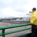 A man in a rain jacket points down at a channelized river below a bridge, full after recent rains.