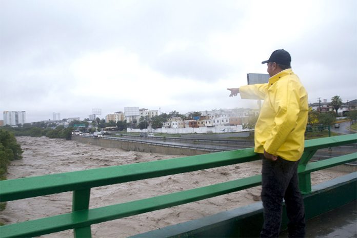 A man in a rain jacket points down at a channelized river below a bridge, full after recent rains.