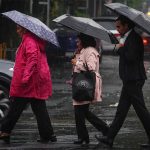 Three Mexico City residents in single file, crossing a rainy street carrying umbrellas.