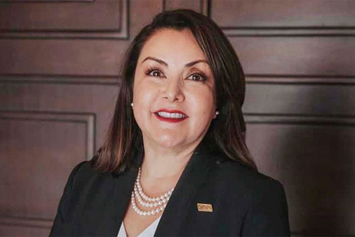 Minerva Pérez Castro in a suit and wearing a pearl necklace, posing in front of a dark-stained wood-paneled wall.