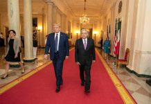 AMLOAMLO and Donald Trump walk down a red carpet in an elegant hallway. and Donald Trump walk down a red carpet in a long corridor.