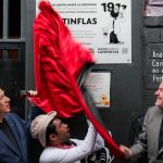 Two older men with a man dressed as Cantinflas pull down a red silk fabric covering a plaque on a building where the iconic Mexican actor grew up.