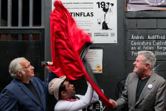 Two older men with a man dressed as Cantinflas pull down a red silk fabric covering a plaque on a building where the iconic Mexican actor grew up.