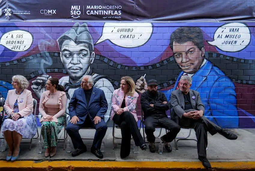 Six people in folding chairs seated in front of a new mural with images of Cantinflas in his most iconic roles in Mexican film