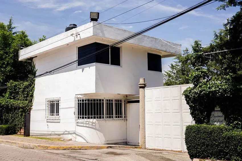 White adobe two-story home with a tall gate on one side and anti-theft metal bars on the first-floor windows.