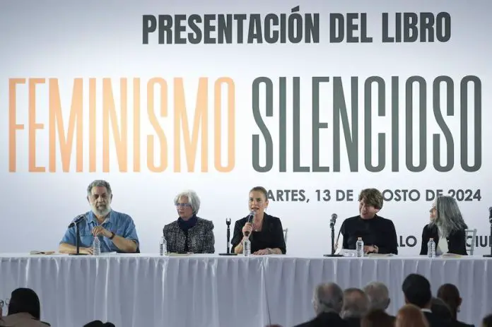 A long conference panel table on a stage with a banquet tablecloth at which are seated five men and women, with scholar and first lady of Mexico Beatriz Gutierrez Muller at the center, speaking into a microphone.