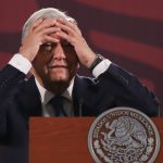 President Lopez Obrador of Mexico standing at a podium at a press conference with his hands on his forehead in a distressed gesture