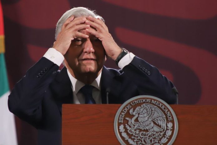 President Lopez Obrador of Mexico standing at a podium at a press conference with his hands on his forehead in a distressed gesture