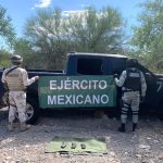 Two soldiers holding up a Mexican army banner by a pickup truck