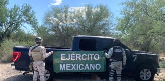 Two soldiers holding up a Mexican army banner by a pickup truck