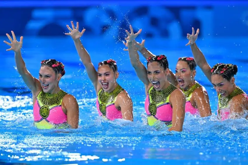 Mexico's synchronized swimming Olympic team in a pool at the Paris Olympics in 2024, each holding a single hand up in the air as part of their routine.