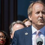 Ken Paxton in a suit and a mauve tie speaking at an event, standing in front of a podium with people watching in the background