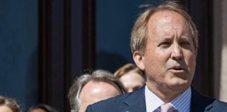Ken Paxton in a suit and a mauve tie speaking at an event, standing in front of a podium with people watching in the background