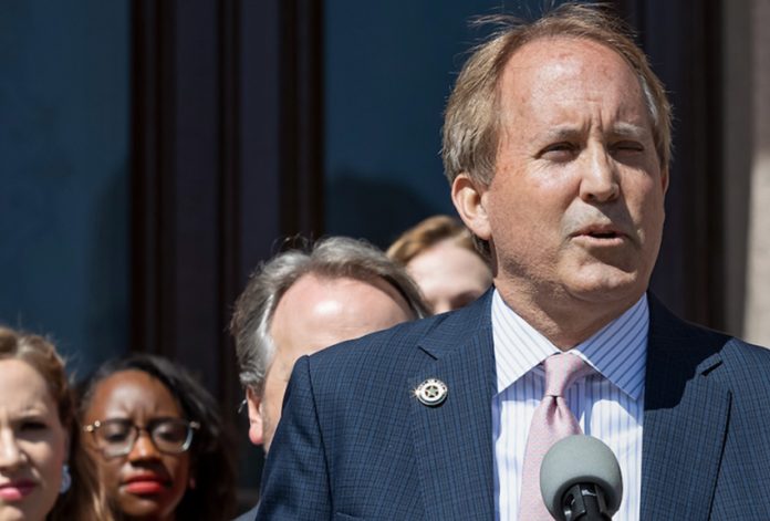 Ken Paxton in a suit and a mauve tie speaking at an event, standing in front of a podium with people watching in the background