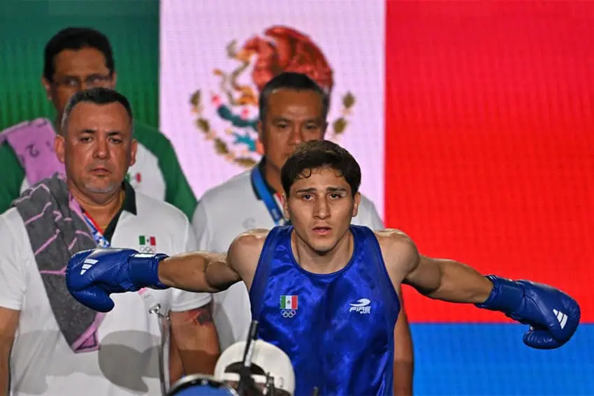 El boxeador Marco Verde de pie con los brazos extendidos frente a los entrenadores con una bandera mexicana al fondo.