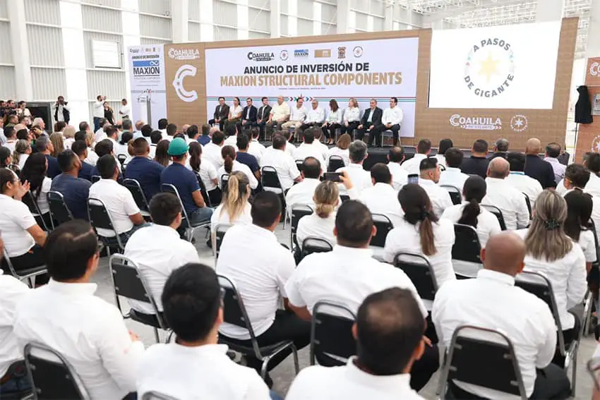 Maxion Structural Components announcement of plant expansion in Monclova, Coahuila. About 100 employees sit in chairs in an audience watching about 12 people seated on a makeshift stage with a banner above them