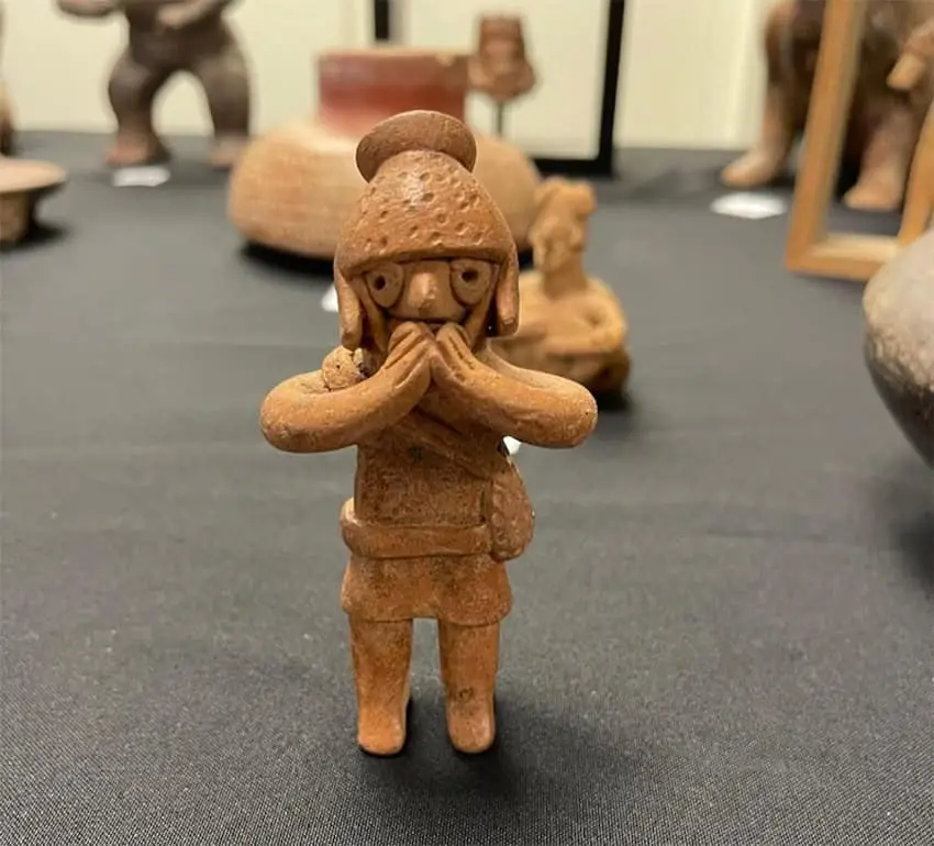 A clay statuette, one of 32 archaelogical artifacts, wearing a helmet with its hands pressed to its mouth, as if surprised.