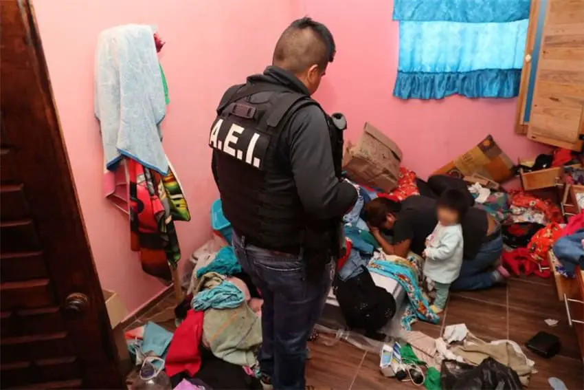 A police officer looks into a messy room, where a young child stands next to a woman kneeling and hiding her face.