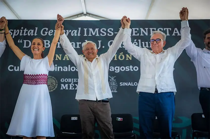 Sinaloa Governor Rubén Rocha Moya stands on stage holding hands with AMLO and President-elect Claudia Sheinbaum.