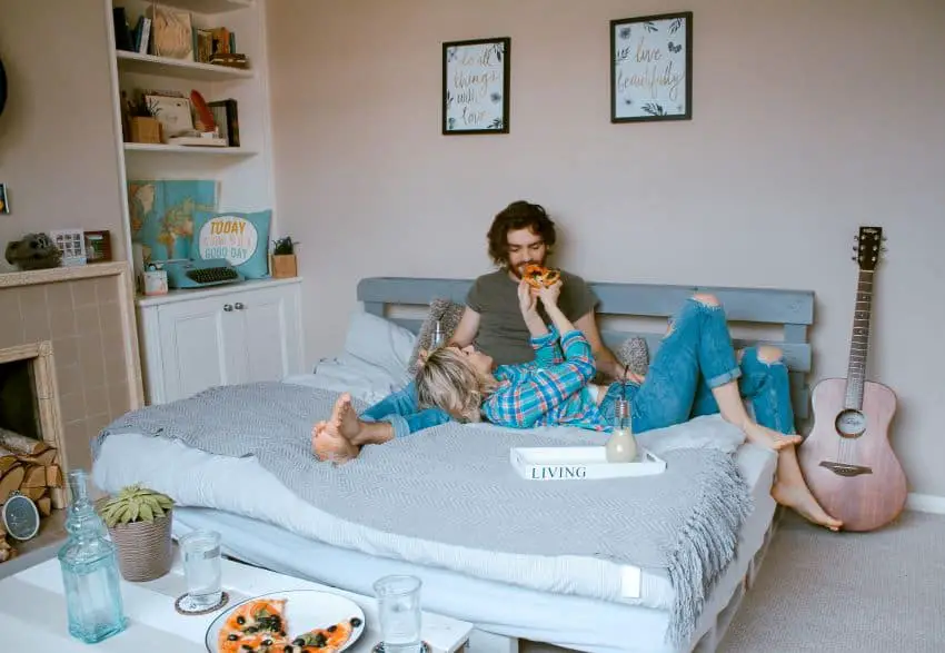 A man sitting up and woman lying down on a bed as the woman feeds the man a slice of pizza.