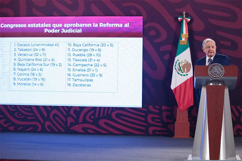 President Lopez Obrador standing at a podium with a projection screen to his right and behind listing all the state legislatures that had already voted to ratify his judicial reform bill.