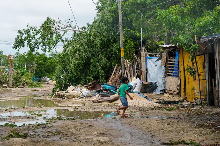 Un niño con una camiseta verde y pantalones cortos de mezclilla corre por un camino fangoso y sin pavimentar lleno de escombros y basura cerca de tres chozas improvisadas de madera y metal que sirven como hogares.