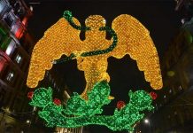 Light mosaic of a yellow eagle with a green serpent in its beak standing on a cactus tree, surrounded nearby by historic buildings in Mexico City's main square