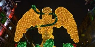Light mosaic of a yellow eagle with a green serpent in its beak standing on a cactus tree, surrounded nearby by historic buildings in Mexico City's main square