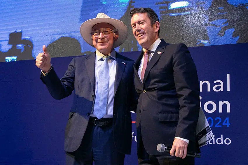 Ken Salazar, wearing a white hat and a dark blue suit and giving a thumbs up as he stands next to Larry Rubin, who holds a microphone. Both are smiling and standing onstage at an American Society of Mexico event where judicial reform was discussed.