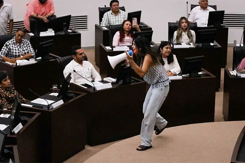 A woman with a megaphone stands in front of seated lawmakers in the Yucatan state congress in session as the woman talks to them and gestures strongly with her hand.