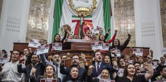 A group of Mexico City legislators standing by the head of the legislature's bench and behind it, holding signs that say "The Reform goes forward!"