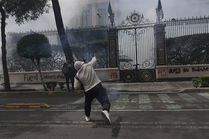 Manifestante con sudadera con capucha, pantalones negros y zapatillas de deporte en la calle frente a la valla frente al edificio de la Secretaría de Gobernación de México. Los muros de piedra de la puerta tienen mensajes políticos en grafitis.