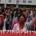 Multiple women standing in rows in a courtyard with their left fists raised in the air. Before the front row is a banner saying "Ayotzinapa, Guerrero."